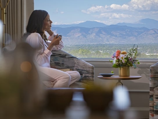 A woman sipping coffee and sitting by her Clio hotel room window, looking at Colorado's mountains; A woman is sitting by the window in her hotel room sipping coffee by her hotel window, looking at Colorado's mountains. On the coffee table are stacked books, a magazine from Clio's Hotel and a vase of flowers.