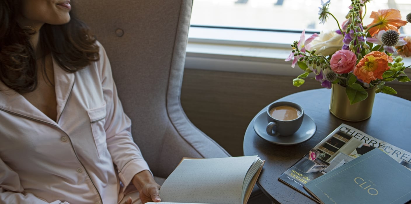 A woman holding a book and drinking coffee in her Clio hotel room; A woman is sitting by the window in her hotel room, holding a book. On the coffee table are stacked books, a magazine from Clio's Hotel, a cup of coffee and a vase of flowers.