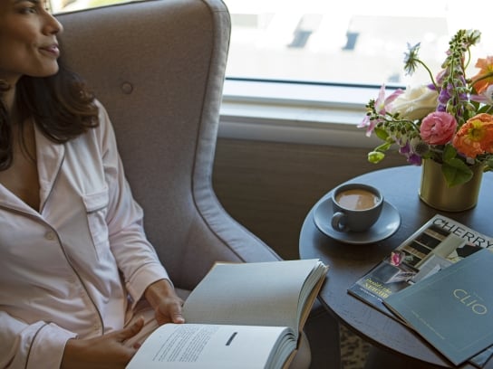 A woman holding a book and drinking coffee in her Clio hotel room; A woman is sitting by the window in her hotel room, holding a book. On the coffee table are stacked books, a magazine from Clio's Hotel, a cup of coffee and a vase of flowers.